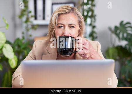 Businesswoman Drinking Coffee While Working on Laptop in a Modern Office Setting Stock Photo