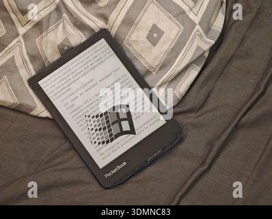 PocketBook Verse Pro ebook reader on bed with grey patterned duvet, e ink screen displaying text about GPT and generative AI with retro Windows 95 log Stock Photo