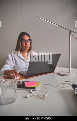 Woman focusing on laptop using a mouse on her white desk with a lamp Stock Photo