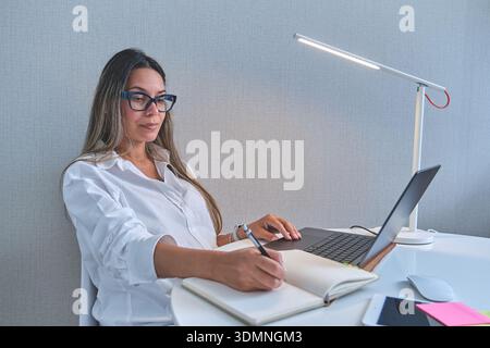 Woman writing notes in notebook while using laptop at her home office Stock Photo