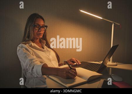Woman writing notes and typing on laptop at desk under lamp light Stock Photo
