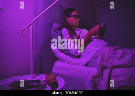 Pregnant woman lying in bed, focused on her smartphone screen in a dark room Stock Photo