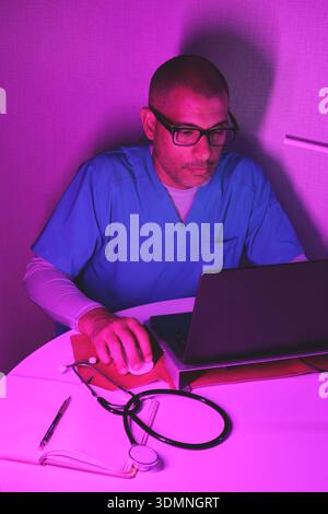Healthcare professional researching medical information on a laptop in a neon-lit room Stock Photo