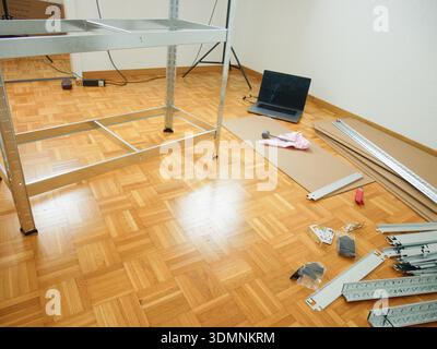 Assembly area featuring metal shelving unit parts, tools, and a laptop on a wooden floor, with a bright indoor environment visible Stock Photo