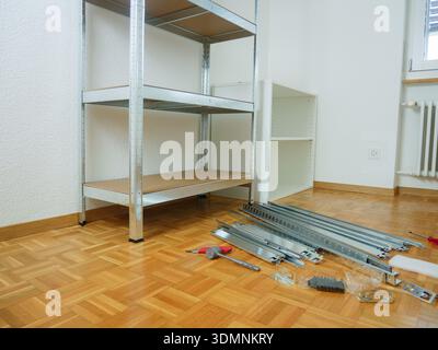 Metal shelving unit is partially assembled in a room with a wooden floor, tools and materials scattered on the ground, and an empty white shelf nearby Stock Photo