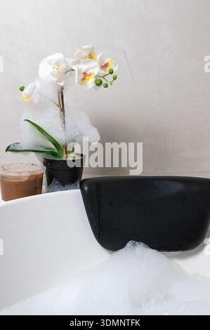 Bathtub bubbles white orchid spa mood, soft foam spilling over tub, black bath pillow draped on rim, potted phalaenopsis beside warm coffee glass, mut Stock Photo