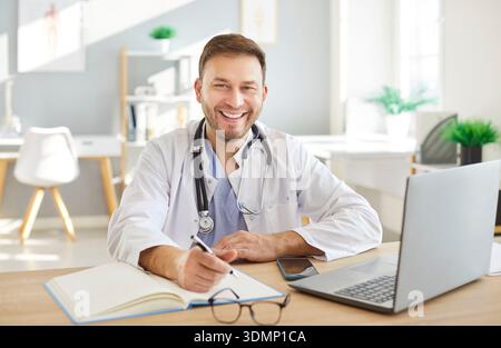 Happy smiling male doctor in white uniform working online on computer, learning, improving  Stock Photo