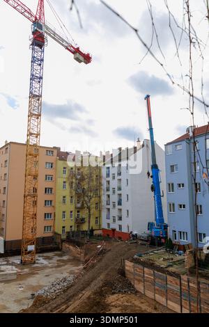 A busy construction yard in a city block, surrounded by pastel apartment buildings. Tall cranes command the sky as workers excavate and lay foundation Stock Photo