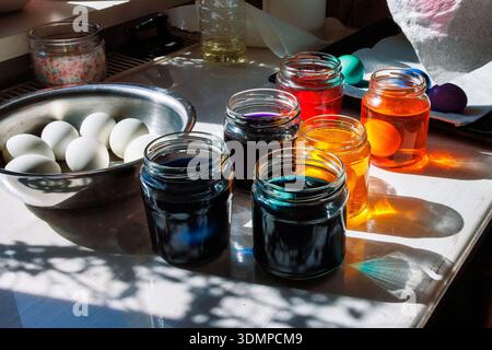 Wide view of a kitchen table set up for easter egg dyeing with a bowl of white eggs and multiple colorful glass jars Stock Photo