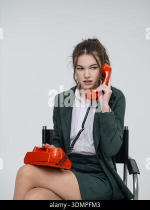 An elegant white woman in a green business suit talks on a red telephone while sitting on a chair against the backdrop of an isolated space. Office. Curly hair, brown-haired Stock Photo
