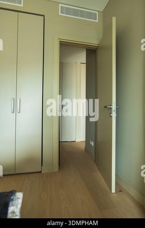 Pale beige walls and light wood flooring frame an open door to a corridor with sleek flush closets, metal handles, and a linear air vent, in a Lithuan Stock Photo
