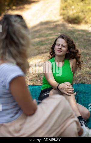 Two women engage in a deep conversation while resting on a green blanket amid trees and grass at a campsite during the day. They enjoy each other's Stock Photo
