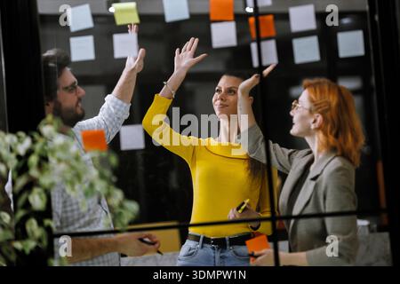 Team brainstorming session in a bright office with colorful sticky notes on a glass wall, showcasing collaboration and creativity during a morning Stock Photo
