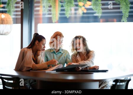 Three businesswomen reviewing documents at a bright table during the day in a modern office space, collaborating on a project and discussing ideas Stock Photo