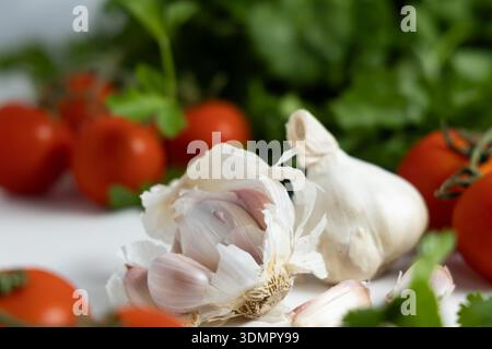 Fresh garlic cloves rest near ripe tomatoes and green herbs on a white surface. Cooking ingredients are ready for use. Stock Photo