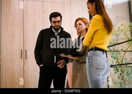 Team members discussing project details while using a laptop in a modern office setting during daytime Stock Photo