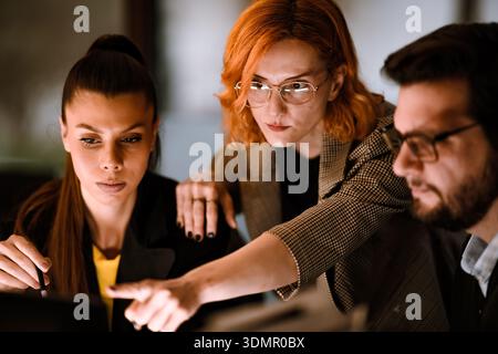Team members collaborate in a modern office space while discussing project details and analyzing data on a computer screen in an evening setting Stock Photo