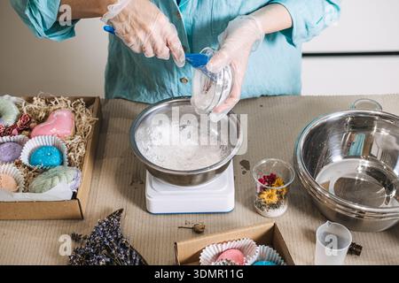 Person pours white powder into stainless steel bowl on digital scale surrounded by colorful finished products. Handmade bath bombs, personalized self Stock Photo