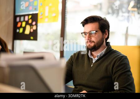 Young professional working in a modern office environment, focused on a task at his desk during daylight hours Stock Photo