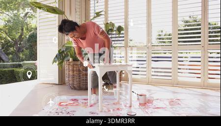 Sanding woman wearing pink top working on small white table amid paint cans and AR overlays Stock Photo