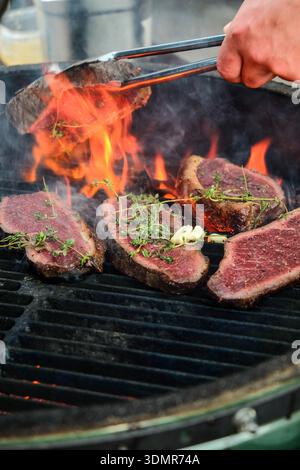 Dynamic close-up of premium beef steaks grilling over open flames, seasoned with fresh herbs and garlic, as a hand turns the meat with metal tongs. In Stock Photo