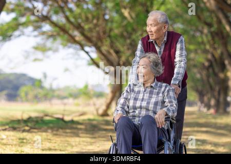 Happy Asian senior couple enjoys park stroll. Elderly man pushes wife in wheelchair Stock Photo