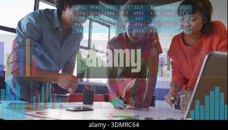 Leaning three team members in business attire analyzing charts on table in office, laptop overlays Stock Photo