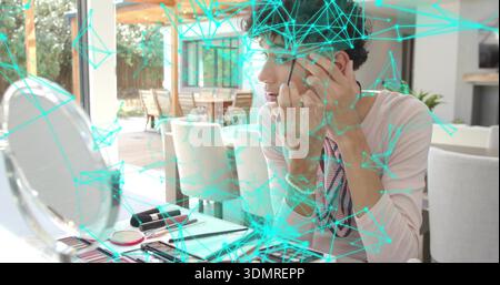Applying eye makeup man wearing light pink top, print scarf at open-plan table, using round mirror Stock Photo