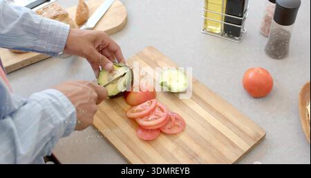 Cutting man in light-blue shirt slicing avocado on wooden board on kitchen counter with tomatoes Stock Photo