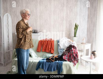 Female fashion enthusiast, dressed in leopard print, stands near a couch with various clothing items, thoughtfully considering her outfit options in a Stock Photo