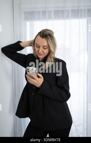 A woman with blonde hair is leaning against a white wall. She is wearing a black blazer and looking down at her phone. There is a white curtain behind her. Stock Photo