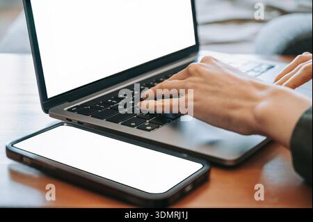 Close-up of hands typing on a modern laptop with a smartphone placed on a wooden desk. Both screens are blank, offering copy space. The image represen Stock Photo