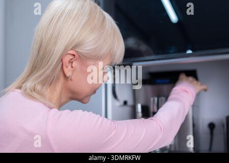 Coffee preparation. Adult blonde woman operating espresso machine holding empty glass cup in bright kitchen interior with focus on routine Stock Photo