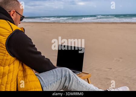 Individual composing social media posts beside ocean. Relaxed author crafting blog content while seated on beach in casual attire Stock Photo