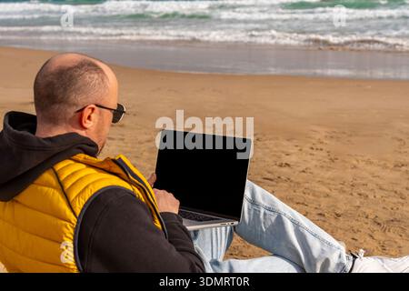 Man working on blog and visuals at sunny shoreline. Casual content editing on laptop amidst beach mood and bright sunlight Stock Photo