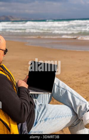 Individual typing on laptop amidst tranquil seaside scenery. Person engaged in coding activities on laptop near peaceful ocean waves Stock Photo