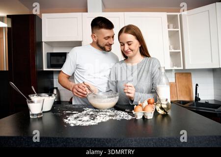 A couple stands in a kitchen working together on a recipe. They mix batter in a bowl while some flour and eggs are on the counter. They enjoy their ti Stock Photo