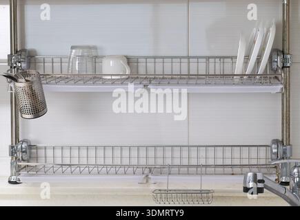 Wall-mounted dish drying rack with plates, glasses and kitchen tools above the sink Stock Photo