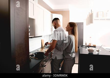 A couple stands close together in a kitchen while one person cooks on the stove and the other hugs them from behind. They enjoy their time cooking dur Stock Photo
