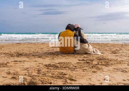 Relaxed pair sitting on shore watching ocean waves. Tranquil scene of partners resting on sandy beach under overcast sky Stock Photo