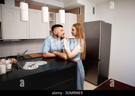 A young couple stands in a kitchen, engaging with each other. The man leans on a black counter while the woman touches his face. They appear to be enj Stock Photo