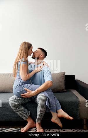A couple is sitting on a couch in a living room. The woman leans in for a kiss with the man. They seem focused on each other and are dressed casually. Stock Photo