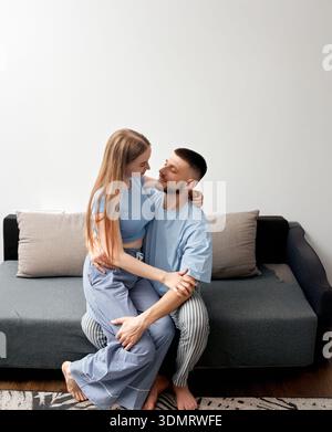 Two people are sitting on a couch in a living room. They are facing each other and looking happy. The light streams in through a window, creating a wa Stock Photo