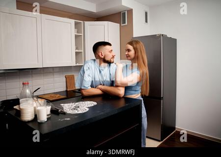 Two people stand close in a kitchen, enjoying each others company. It appears to be afternoon as they smile and engage in light conversation. Stock Photo