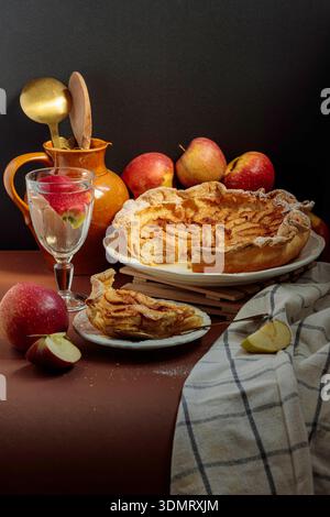 Autumn still life with apple pie, fresh apples and ceramic jug on brown table. Cozy rustic dessert composition. Stock Photo