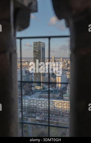 High vantage view of Warsaw, Poland, featuring a modern skyscraper, urban buildings, and a window frame with metal bars under a cloudy sky. Stock Photo