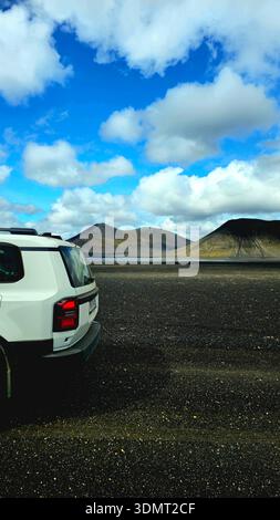 A white SUV sits on a dark volcanic plain under a blue sky with clouds. Two barren mountains with earthy tones rise in the background. Stock Photo