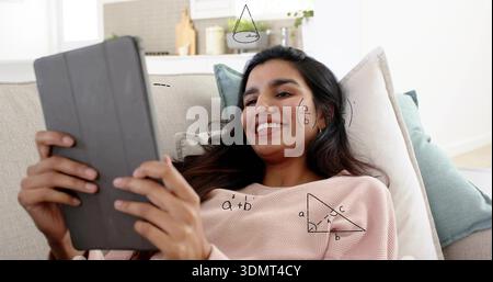 Reclining Indian woman wearing pink sweater holding tablet at home on couch, showing math doodles Stock Photo