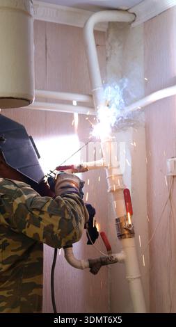 A welder, seen from behind, wearing a uniform and hard hat, welds a section of a house's heating system, made of white-painted iron pipes Stock Photo