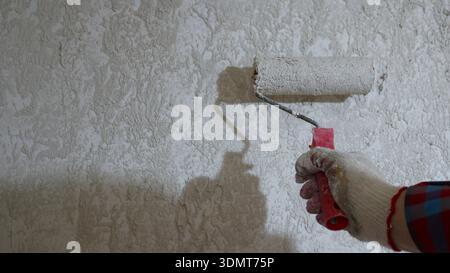 A roller in a gloved hand applies mortar or plaster to a gray wall in a textured, uneven layer, close-up, plastering and preparing the wall surface Stock Photo
